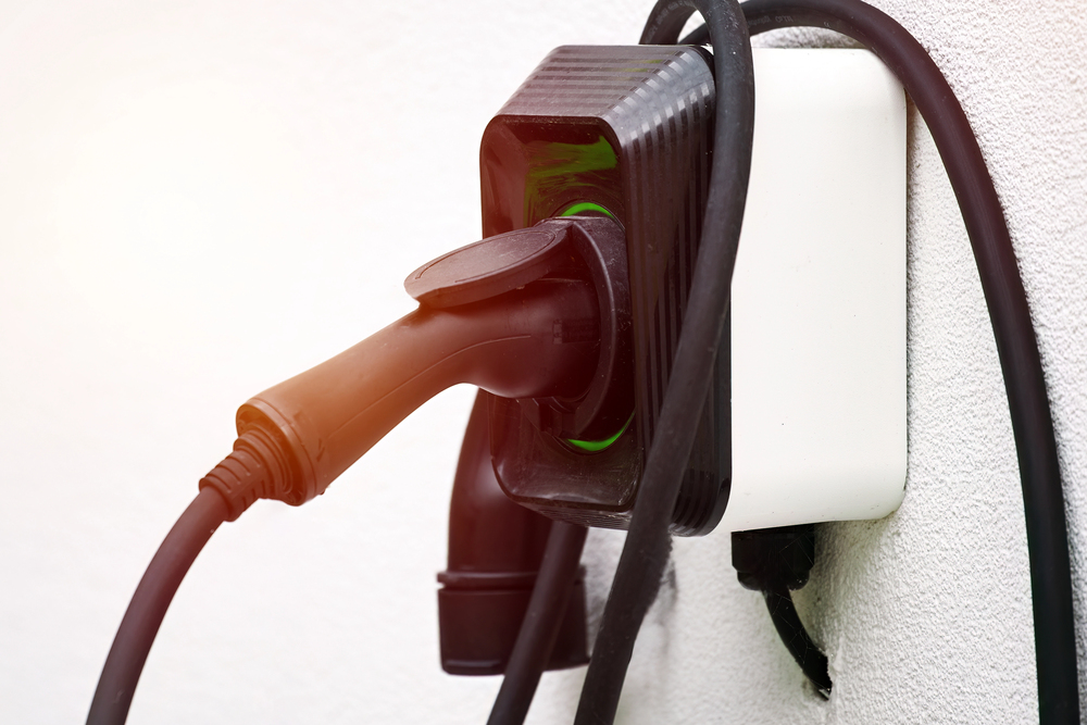 A black electric vehicle charging plug is connected to a wall-mounted charging box, with the cable looped around the unit.