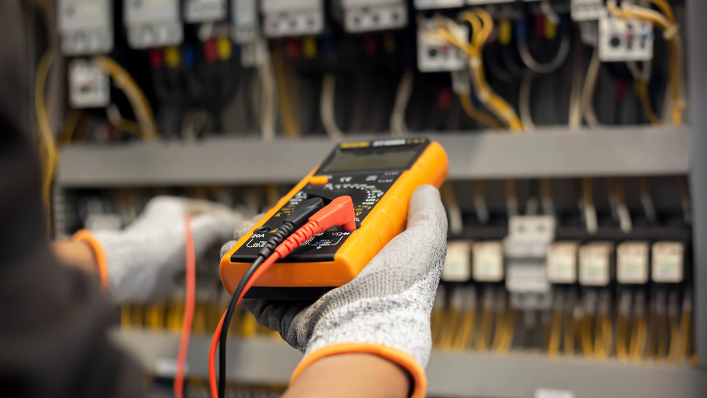 A person wearing protective gloves holds a digital multimeter with test probes in front of an open electrical control panel.