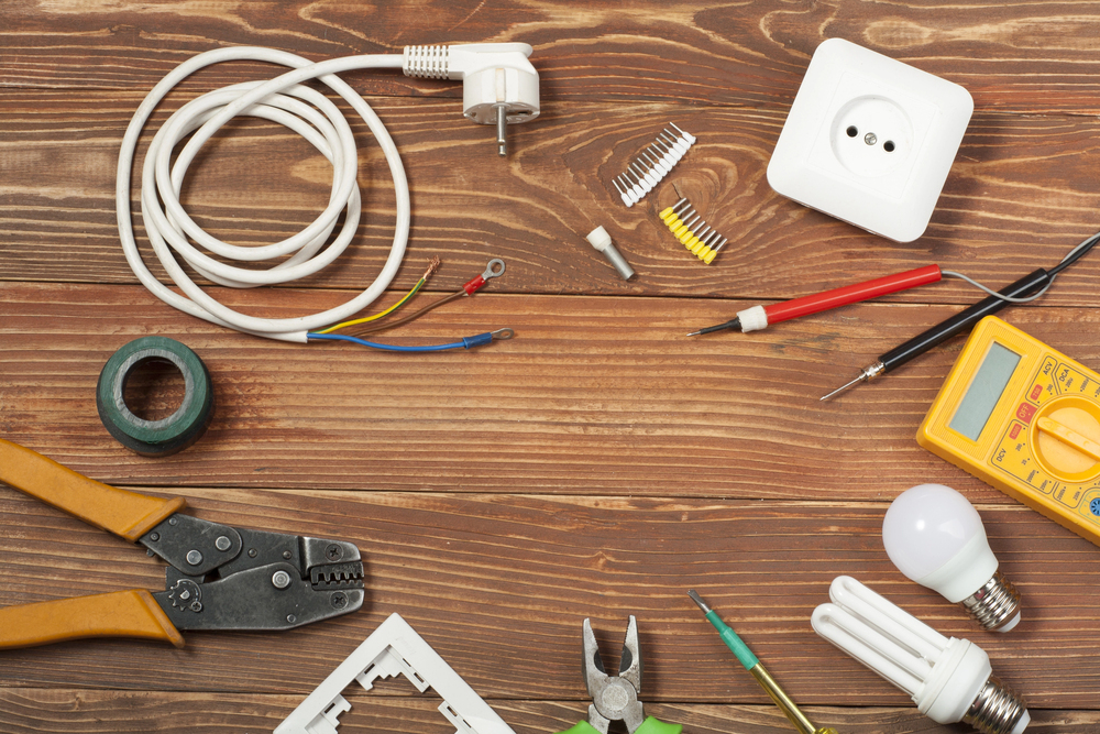 Various electrical tools and components, including wires, pliers, a socket, a multimeter, screwdrivers, tape, and light bulbs, are arranged on a wooden surface.