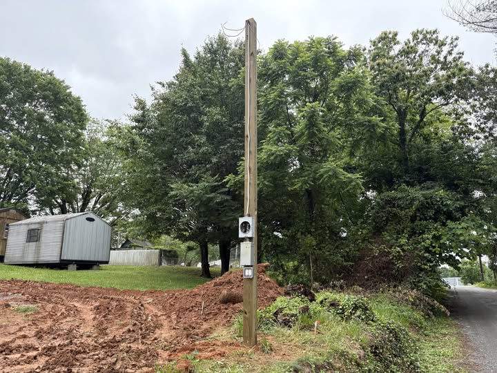 A utility pole with electrical meters stands on a patch of red soil near a dirt path, with trees and a small gray building in the background.