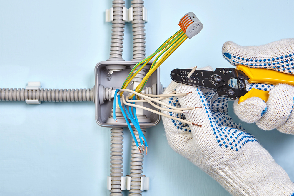 Gloved hands use a wire stripper to strip insulation from electrical wires inside a junction box with several connected conduits.