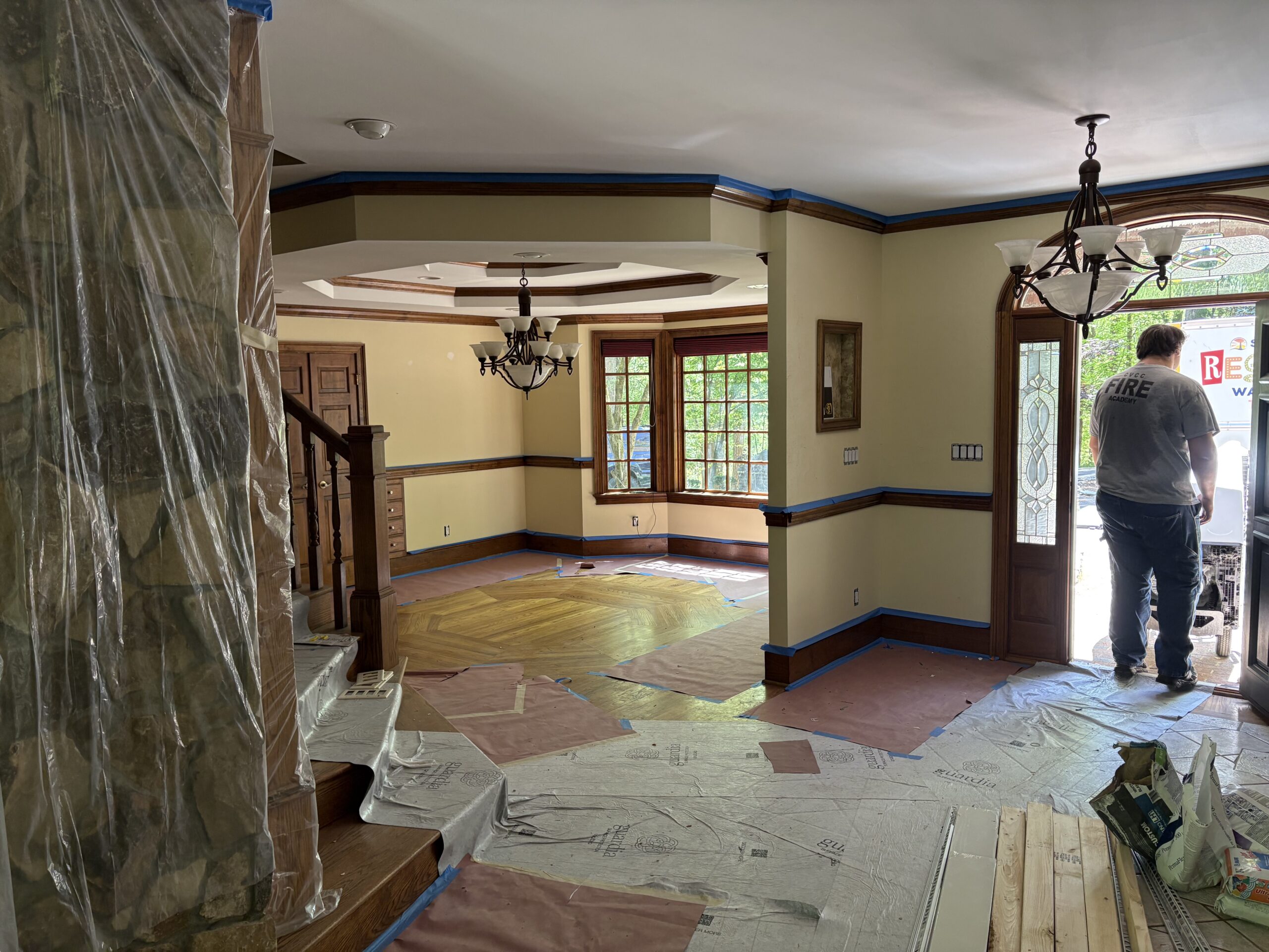 Interior of a house under renovation with protective coverings on the floor and stairs; a person stands at an open door near renovation materials, highlighting ongoing electrical installation services.
