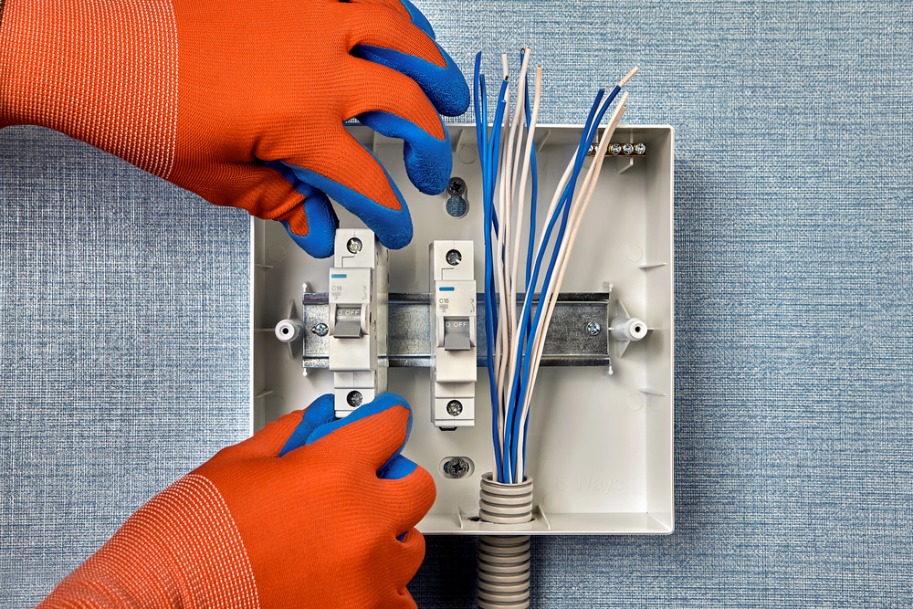 A person wearing orange gloves works with circuit breakers and electrical wiring inside a wall-mounted box.