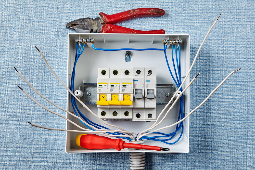 An open electrical junction box with exposed wires, circuit breakers, a red-handled screwdriver, and pliers resting on top, set against a blue textured background.
